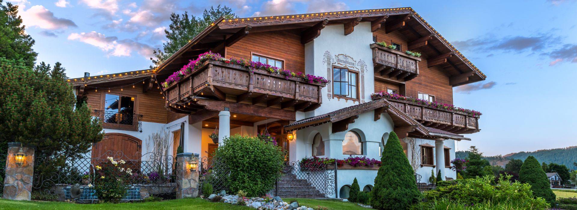 Front facade of a three story white and brown home in old word style wiith balconies lined in flowers, lush landscaping and arched window details