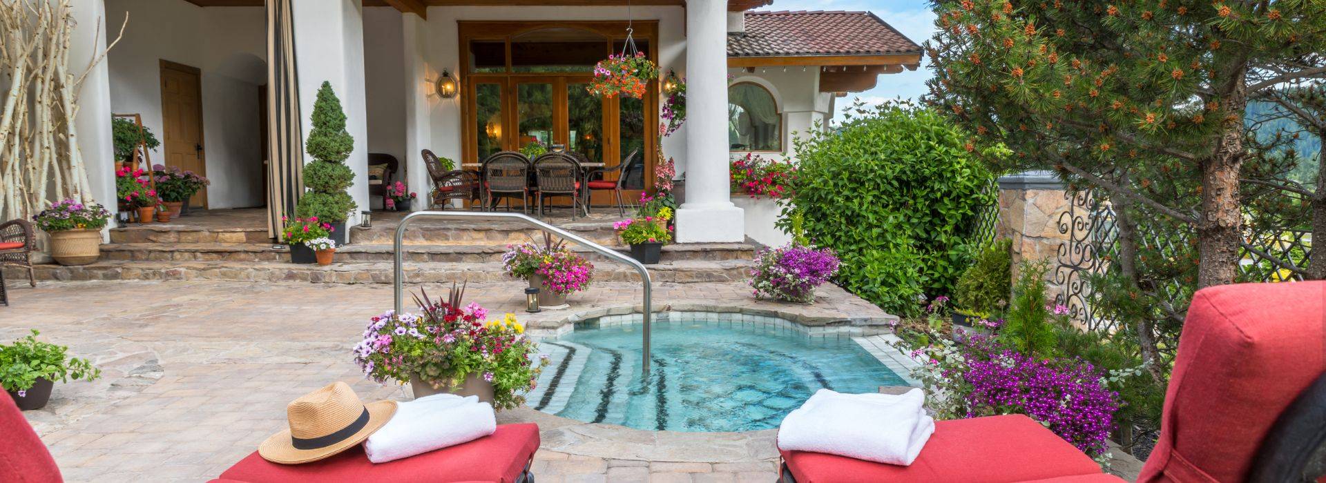Spacious outdoor patio of a luxury two story home with hot tub, red lounging chairs, patio table and chairs and flower pots all over
