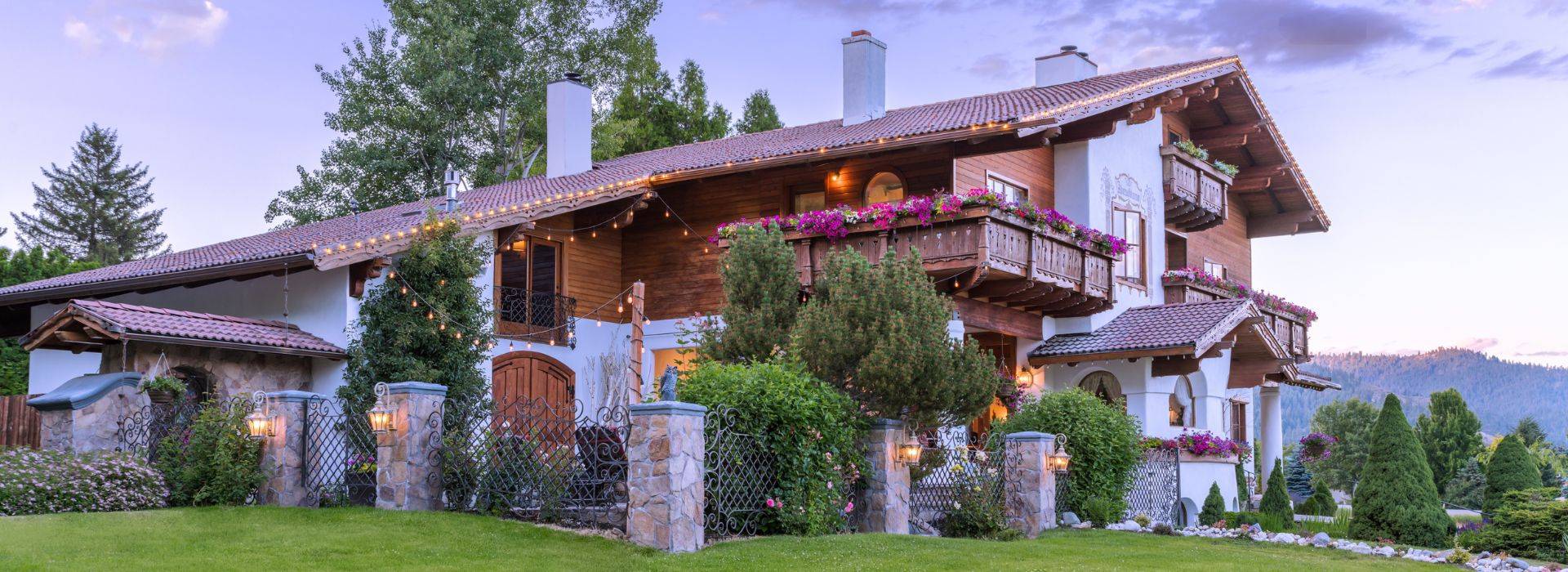 A large, luxurious white and brown home in old world Bavarian style architecture surrounded by trees and lush landscaping with mountains in background